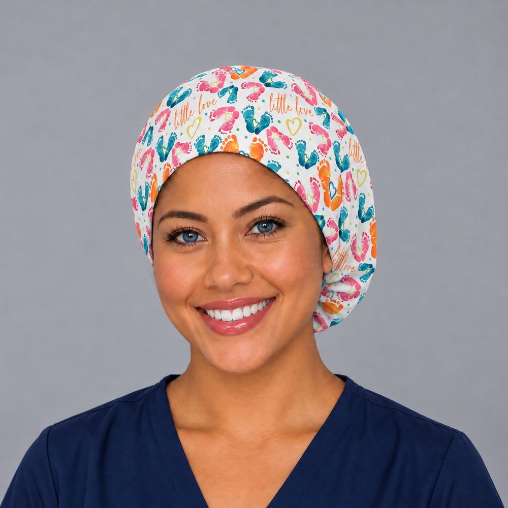 Smiling nurse in navy scrubs wearing a baby feet labor and delivery euro scrub cap, front view with orange "little love" script and footprints across the forehead