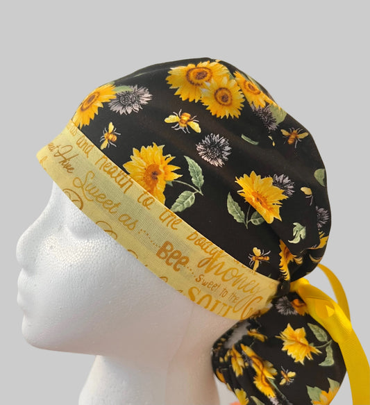 Bee Grateful ponytail scrub cap with sunflowers, bees & yellow script band, front view — handmade-in-USA surgical hat for women with long hair from OR World.