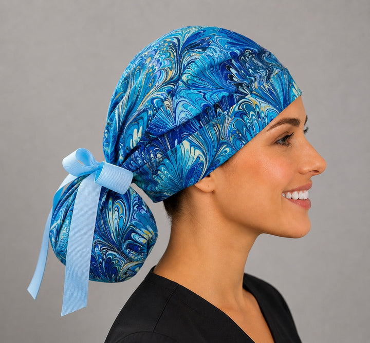 Woman wearing blue marble ocean swirl ponytail scrub cap, front view smiling with blue, turquoise and gold marbled feather print and light blue ribbon tie