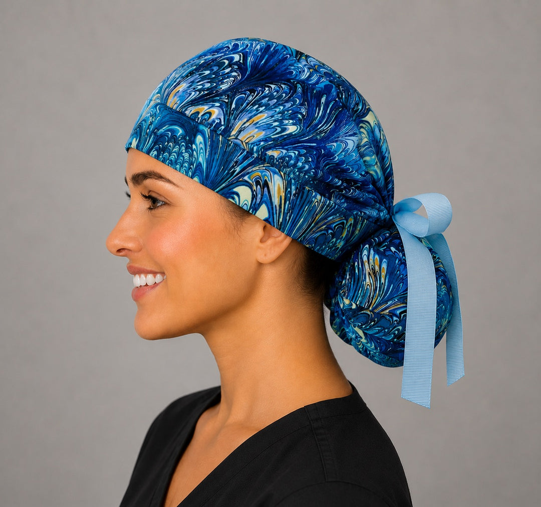 Blue marble ocean swirl ponytail scrub cap left side profile with full ponytail pouch of turquoise, navy and gold marbled feather print and long light blue ribbon ties