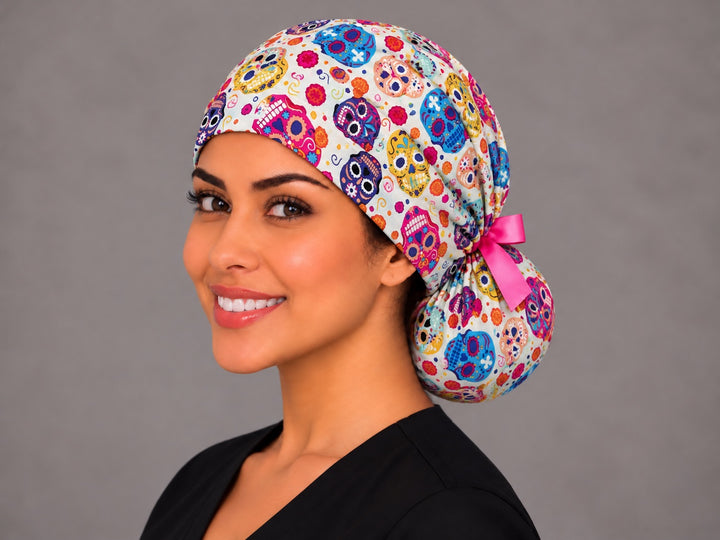 Three-quarter angle of a calavera ponytail nurse hat, with pink, yellow and violet skulls and orange marigolds across the gathered crown