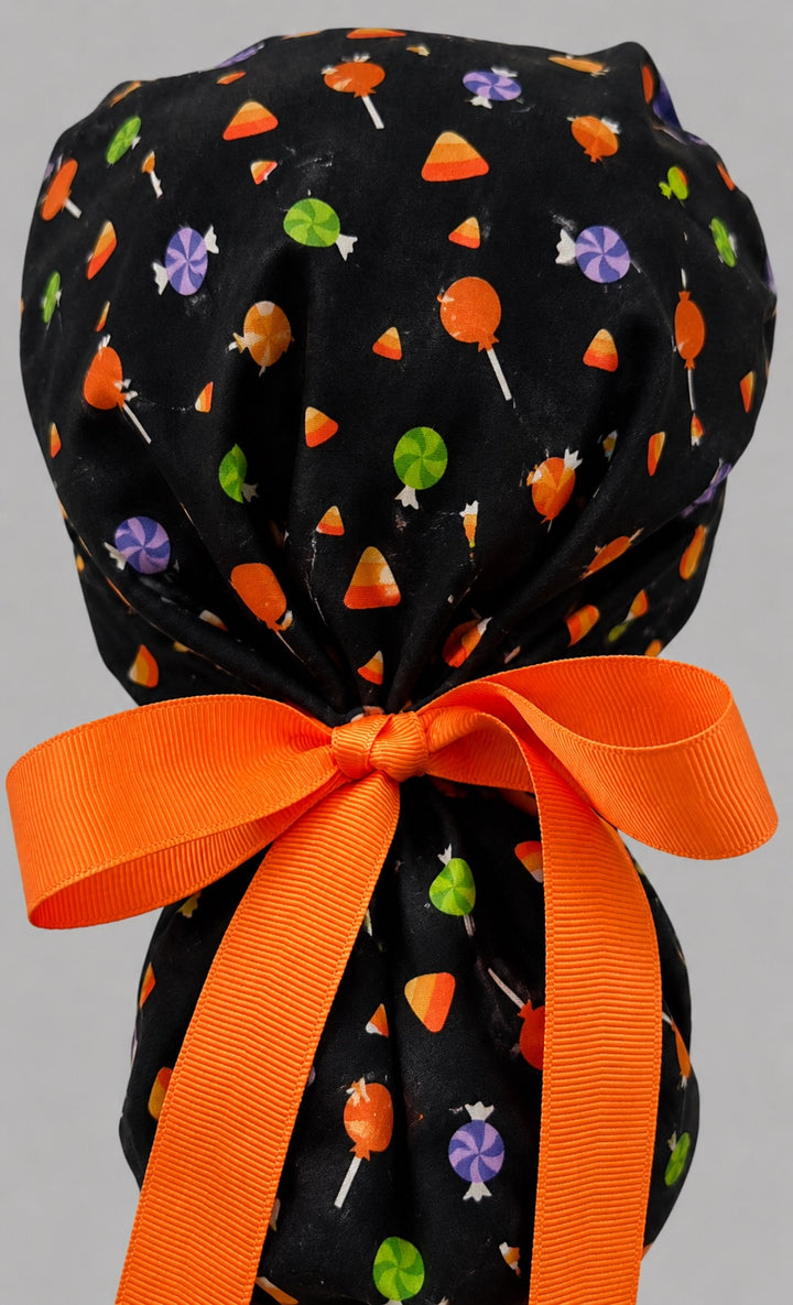 Halloween candy corn ponytail surgical cap left side profile showing candy corn, lollipops and wrapped candy on black with orange ribbon tie for long hair
