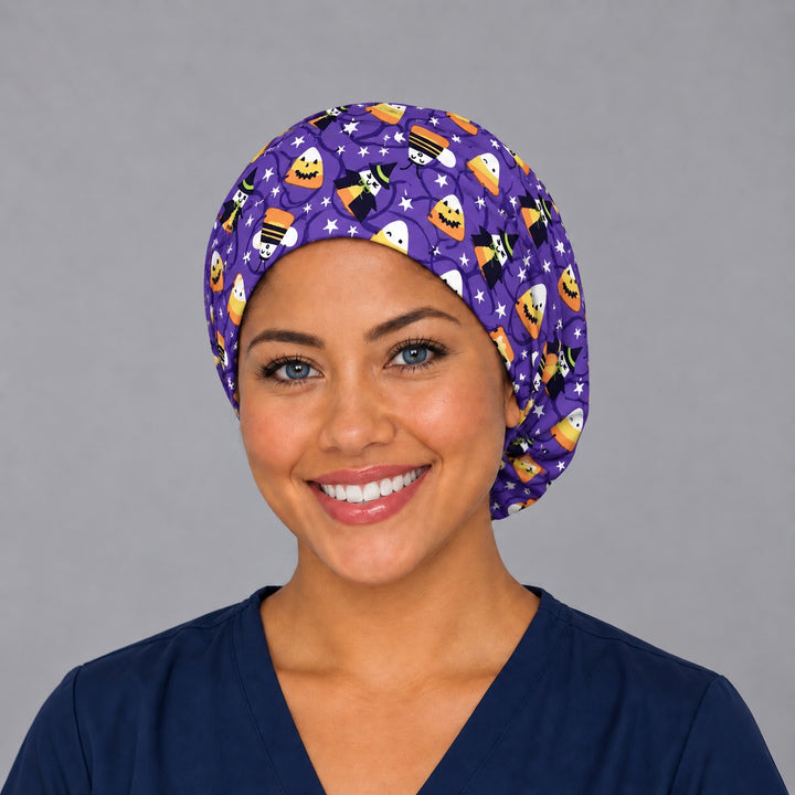 Woman wearing a candy corn witches Euro scrub cap, front view — smiling candy corn and witches on purple, adjustable Halloween surgical hat