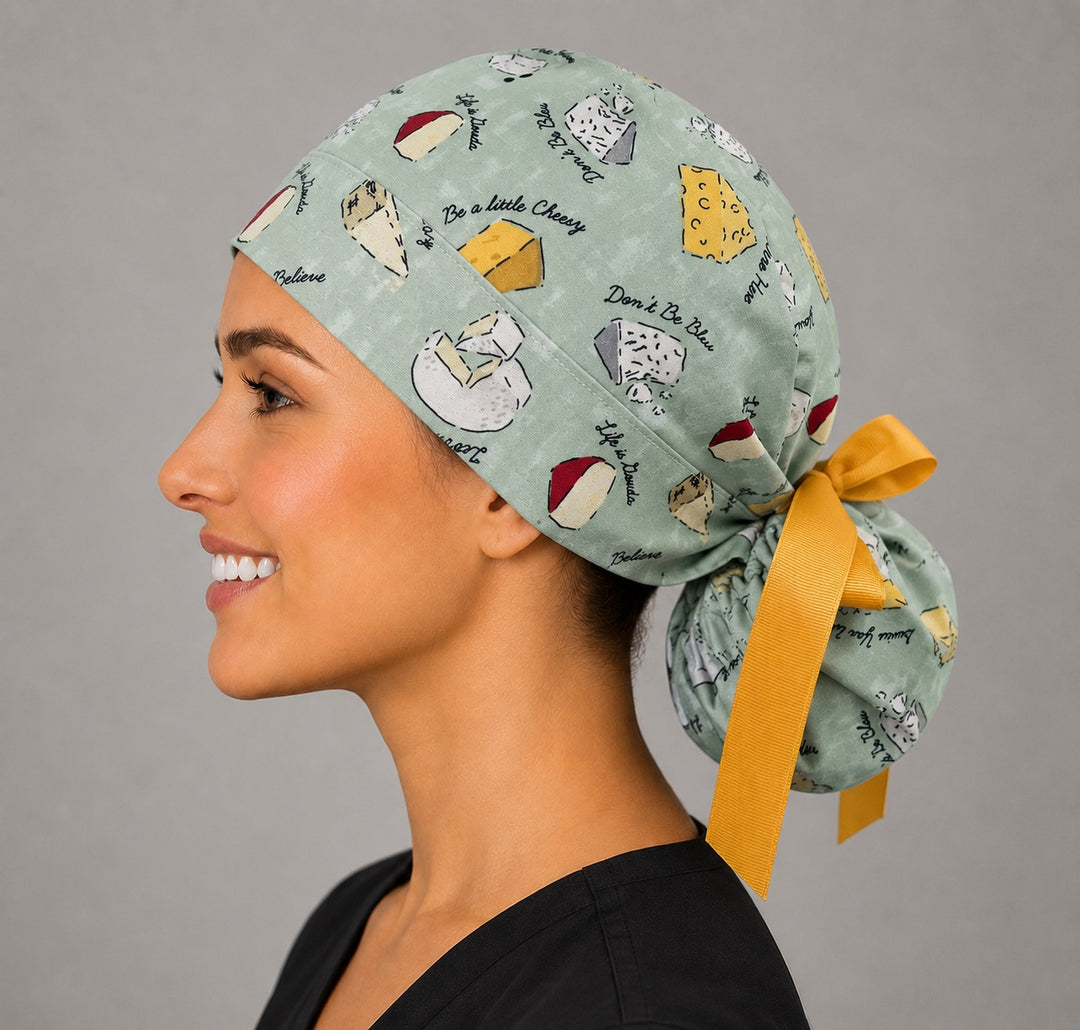 Funny cheese pun ponytail scrub cap left side profile with cheese wedges, Be a Little Cheesy sayings on sage green and yellow grosgrain ribbon tie