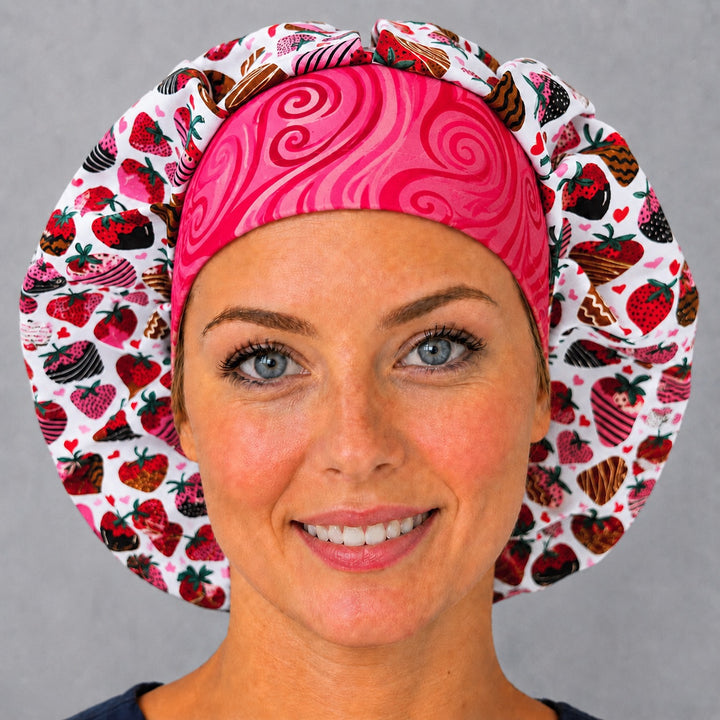 Woman wearing a chocolate-covered strawberry bouffant scrub cap, front view — chocolate-dipped berries and hearts with pink swirl band, adjustable women&