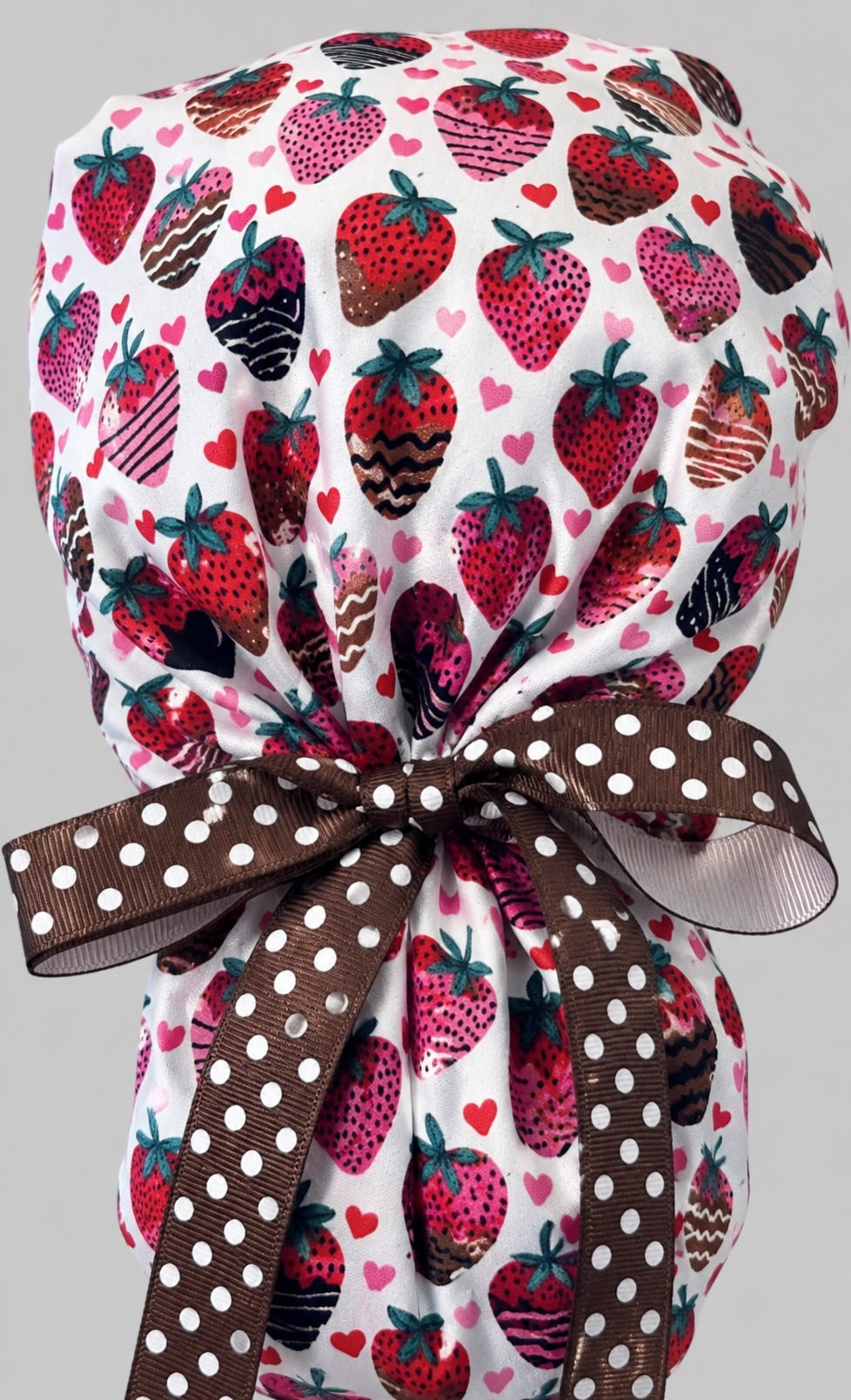 Chocolate-covered strawberry ponytail scrub cap back view close-up showing gathered pouch full of chocolate-dipped strawberries and hearts on white with brown polka-dot ribbon bow