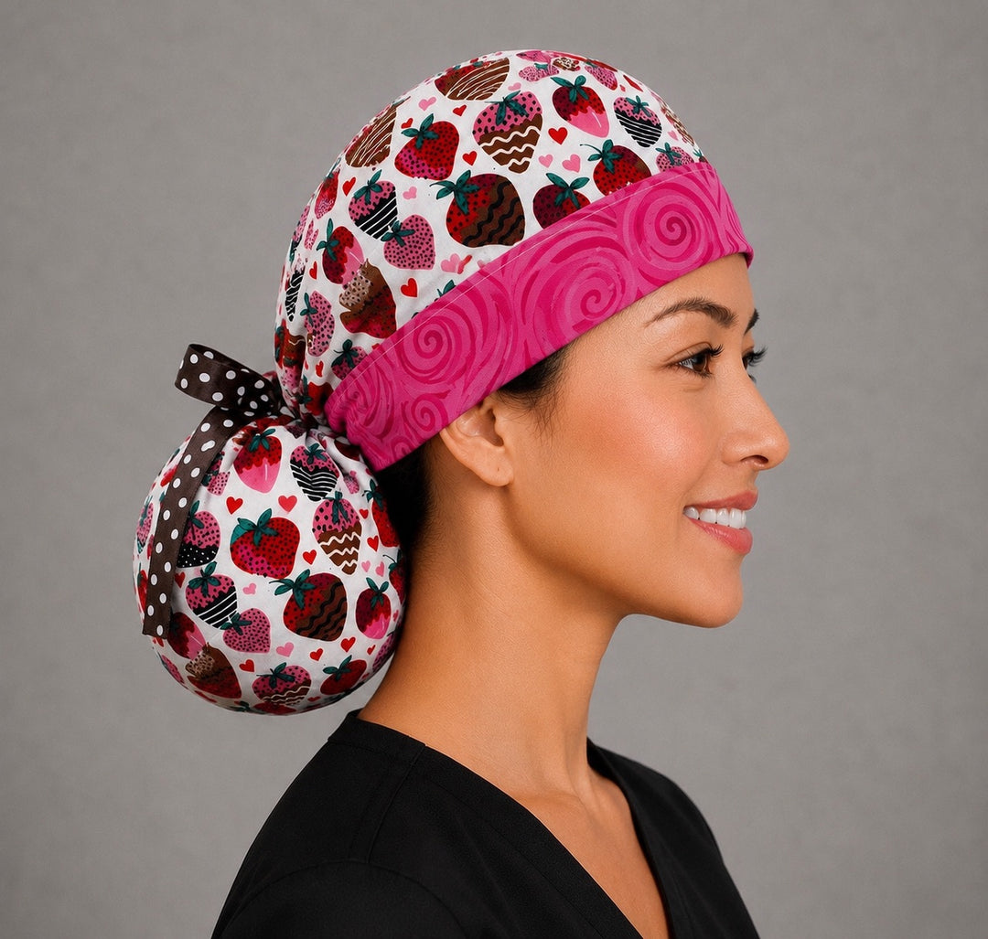 Woman wearing chocolate-covered strawberry ponytail scrub cap, front view smiling with chocolate-dipped strawberries and hearts on white with pink swirl band and brown polka-dot ribbon tie