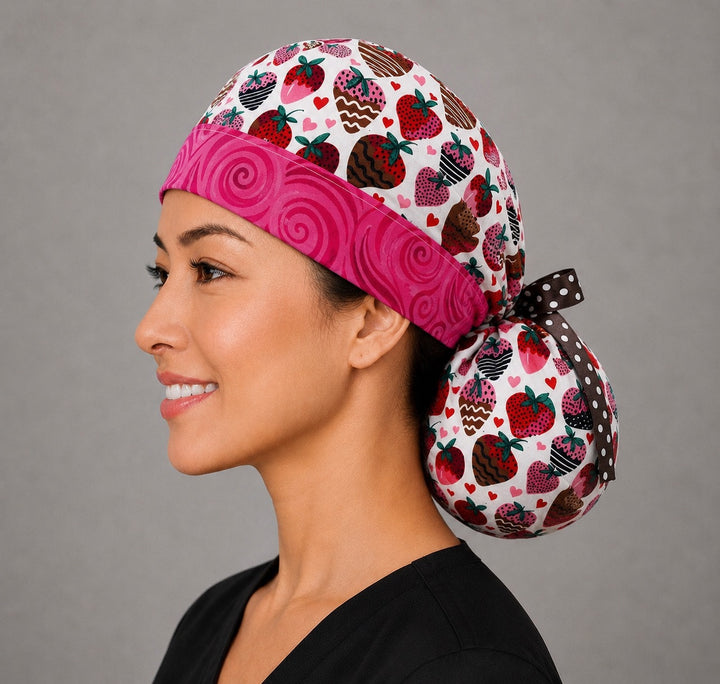Chocolate-covered strawberry ponytail surgical cap left three-quarter angle showing chocolate-dipped strawberries and hearts with pink swirl band and brown polka-dot ribbon bow