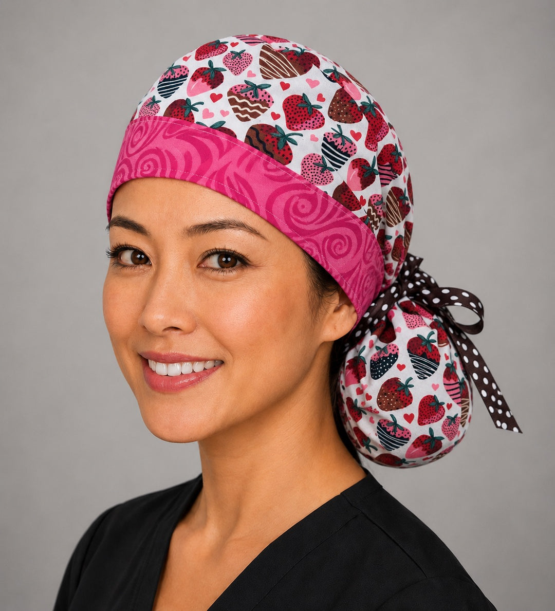 Chocolate-covered strawberry ponytail nurse cap right three-quarter angle on smiling model showing chocolate-dipped strawberries, hearts and pink swirl band with brown polka-dot ribbon bow