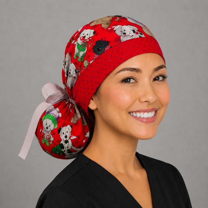 Woman wearing Christmas dogs ponytail scrub cap, front view smiling with pups in Santa hats and sweaters on red with red polka-dot band and white ribbon tie