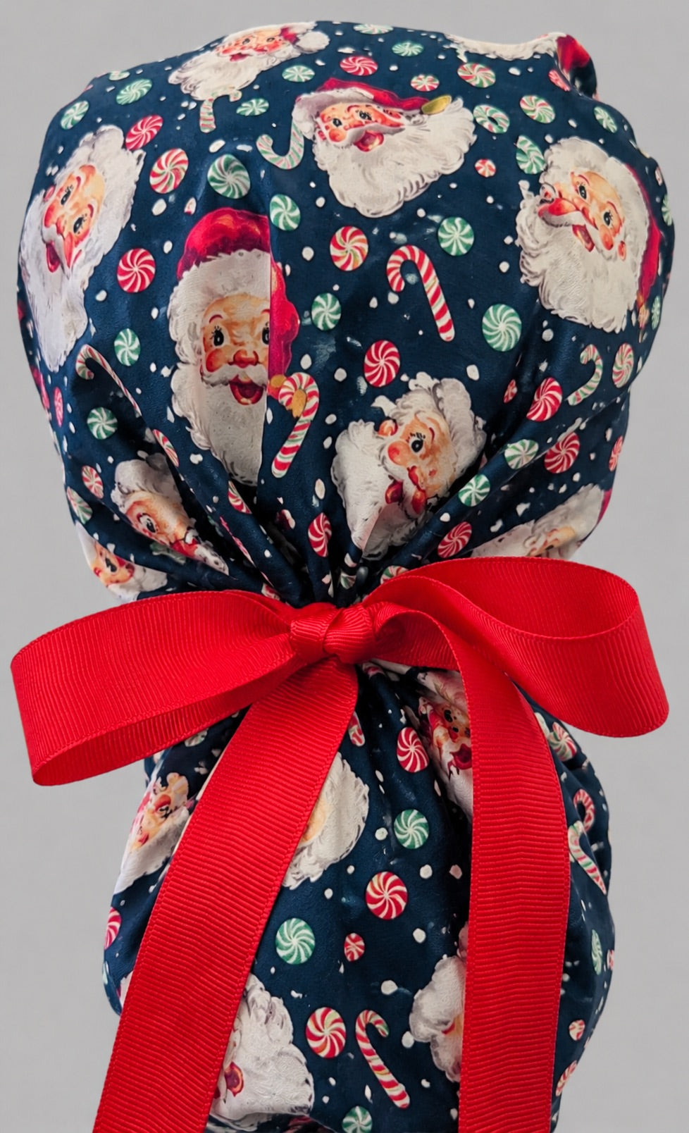 Christmas Santa ponytail scrub cap back view close-up showing gathered pouch full of vintage Santa faces, peppermints and candy canes on navy with red grosgrain ribbon bow