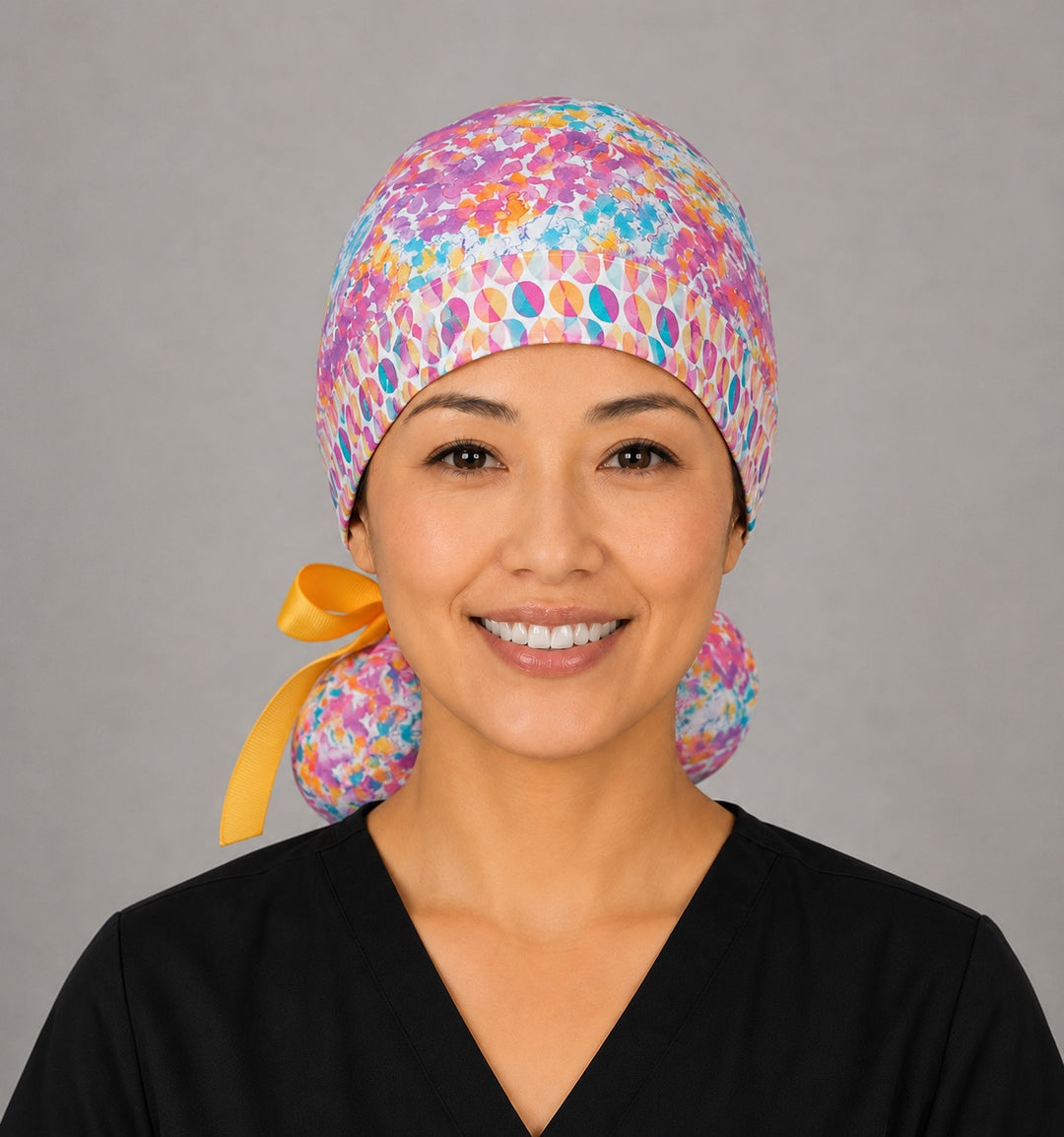 Woman wearing colorful floral watercolor ponytail scrub cap, front view smiling with pink, teal and orange watercolor floral print and gold ribbon tie