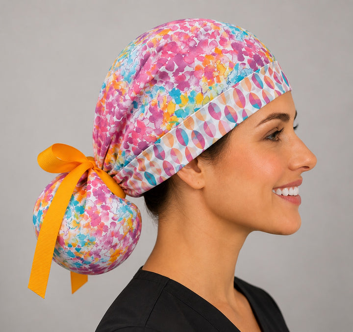Colorful floral watercolor ponytail nurse cap right three-quarter angle on smiling model showing pink teal orange watercolor print with geometric band and gold ribbon bow