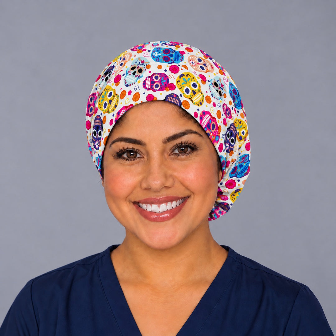 Smiling nurse in navy scrubs wearing a cream sugar skull euro scrub cap, front view with yellow, pink and blue calaveras and orange marigolds across the forehead
