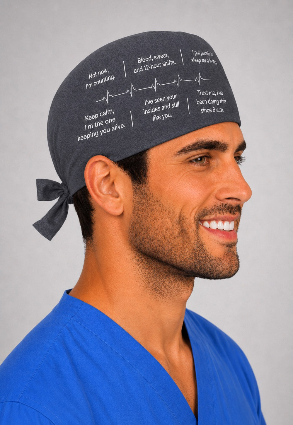 Custom quote scrub cap in charcoal gray with funny personalized medical sayings in white text and a heartbeat line, front view on a smiling male surgeon in blue scrubs, your own words custom text surgical hat, handmade in USA gift for surgeons and nurses, OR World