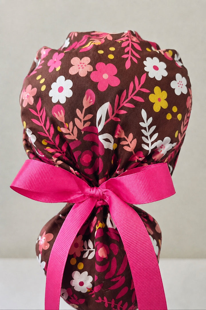 Dark brown floral ponytail scrub cap back close-up showing pink, coral, yellow and white flowers on rich dark brown fabric with hot pink ribbon bow