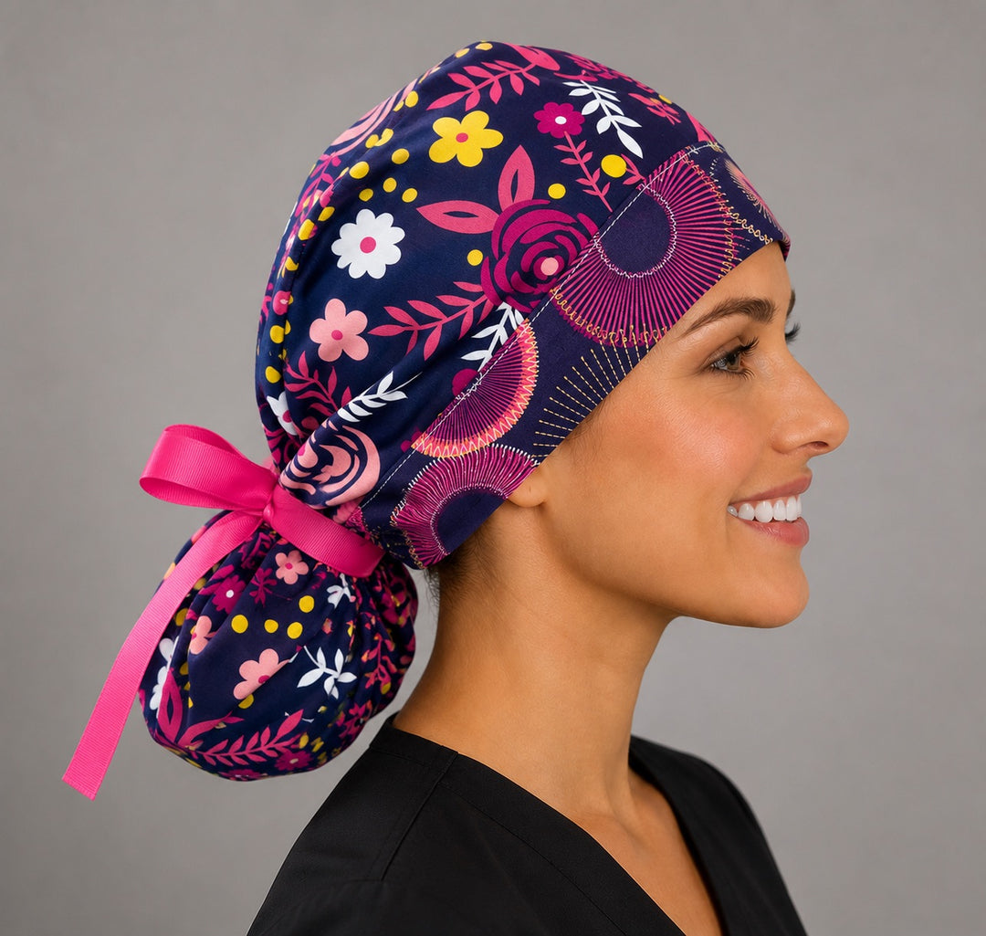 Woman wearing dark brown floral ponytail scrub cap, front view smiling with bright pink, yellow and white flowers on dark brown with pink ribbon tie