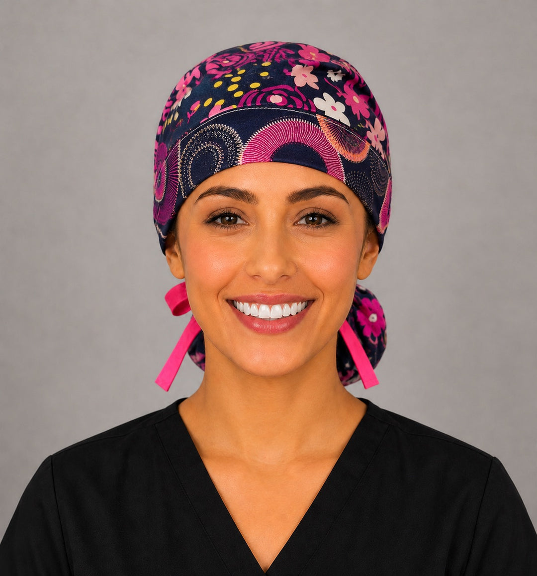 Dark brown floral ponytail nurse cap left three-quarter angle on smiling model showing pink, yellow and white flowers with pink sunburst band and pink ribbon bow