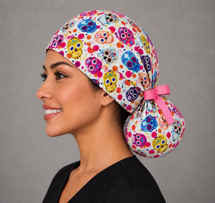 Side profile of a Day of the Dead ponytail surgical hat, showing a short hot pink bow tied above the round ponytail pouch at the nape
