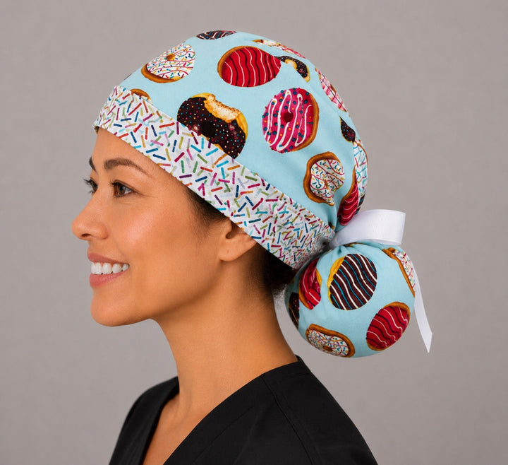 Left side view of donut print ponytail scrub cap for women showing sprinkle headband, roomy ponytail pouch of frosted donuts on aqua and white ribbon bow