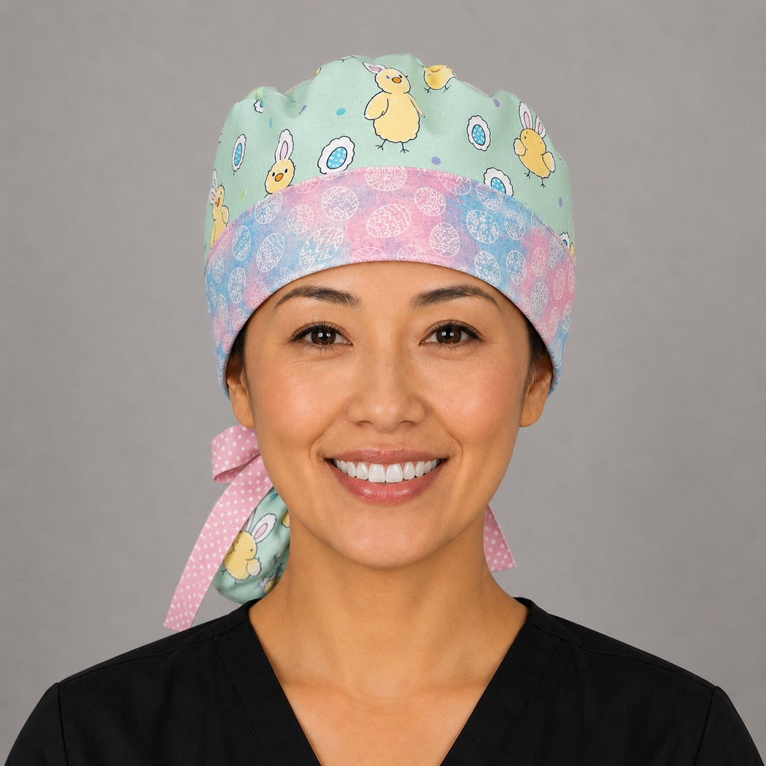 Woman wearing Easter chickens ponytail scrub cap, front view smiling with bunny-ear chicks and Easter eggs on aqua with pastel egg band and pink polka-dot ribbon tie