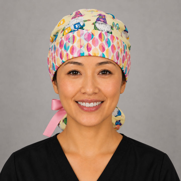 Woman wearing Easter gnomes ponytail scrub cap, front view smiling with pastel gnomes, Easter eggs and baskets on light yellow with egg band and pink ribbon tie