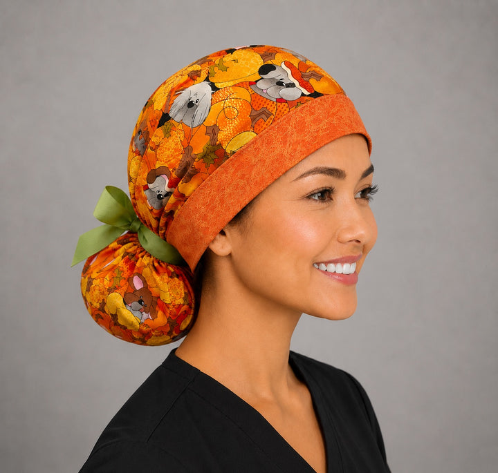 Woman wearing fall dog ponytail scrub cap, front view smiling with puppies among pumpkins and autumn leaves with orange band and green ribbon tie