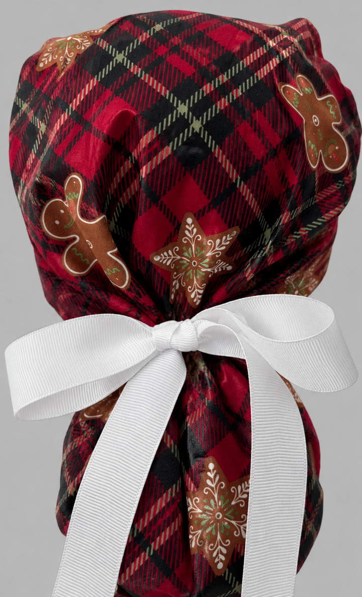 Woman wearing gingerbread treats ponytail scrub cap, front view smiling with red plaid, gingerbread men and star cookies with gingerbread band and white ribbon tie