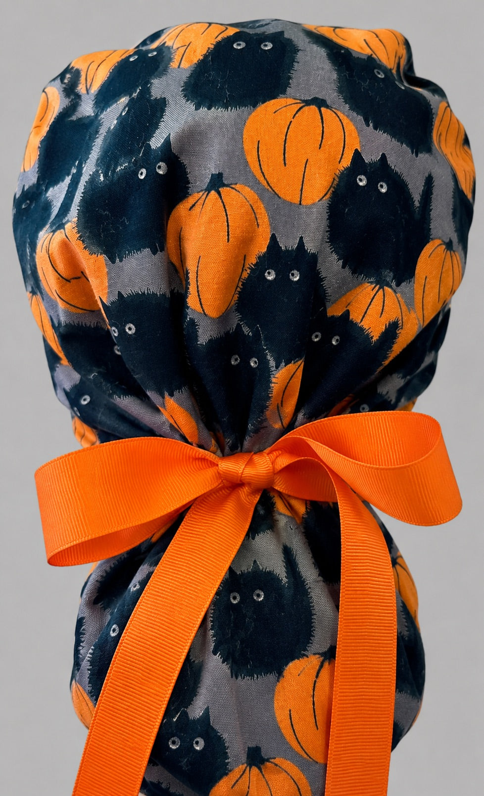 Woman wearing Halloween black cat and pumpkin ponytail scrub cap, front view smiling with fuzzy black cats and orange pumpkins on gray with orange band and orange ribbon tie