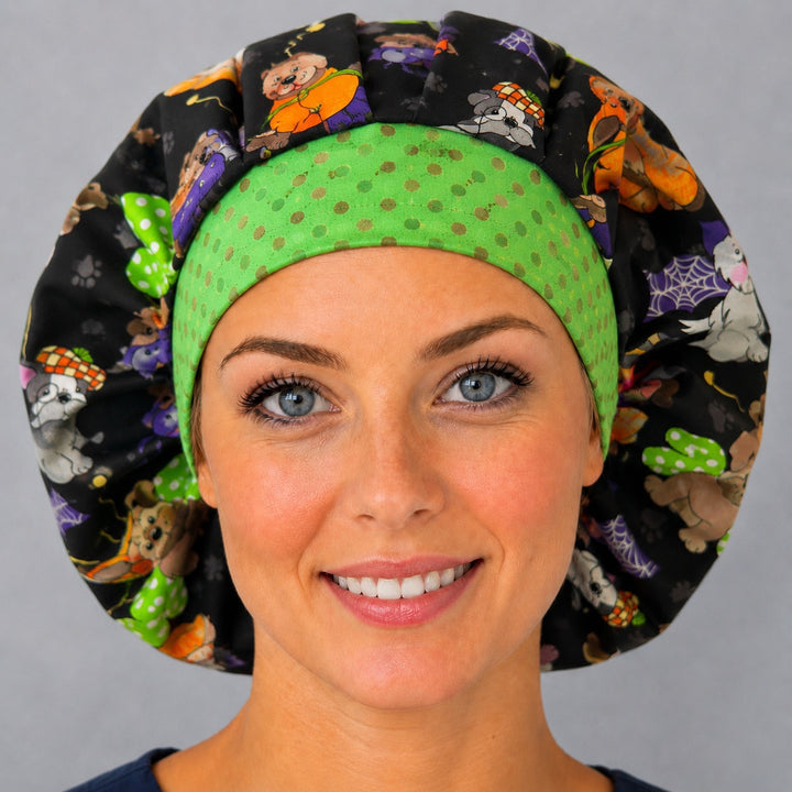 Woman wearing a Halloween dog bouffant scrub cap, front view — costumed pups on black with a green polka-dot band, adjustable women&