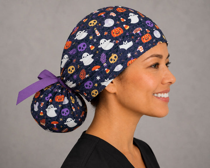 Cute ghost and pumpkin Halloween ponytail scrub cap back view close-up showing gathered pouch full of ghosts, smiling pumpkins, skulls and candy corn on starry navy with purple ribbon bow
