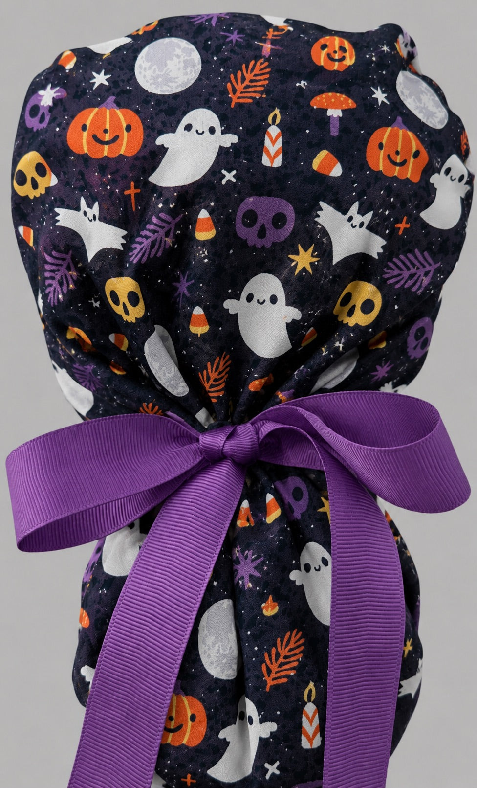 Woman wearing cute ghost and pumpkin Halloween ponytail scrub cap, front view smiling with ghosts, pumpkins, skulls and candy corn on navy with purple ribbon tie