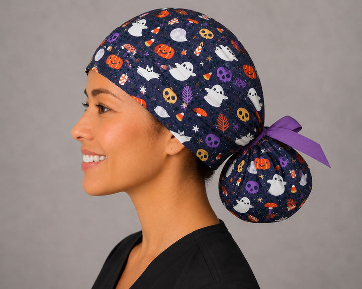 Cute ghost and pumpkin Halloween ponytail nurse cap right three-quarter angle on smiling model showing ghosts, pumpkins, candy corn and skulls on navy with purple ribbon bow