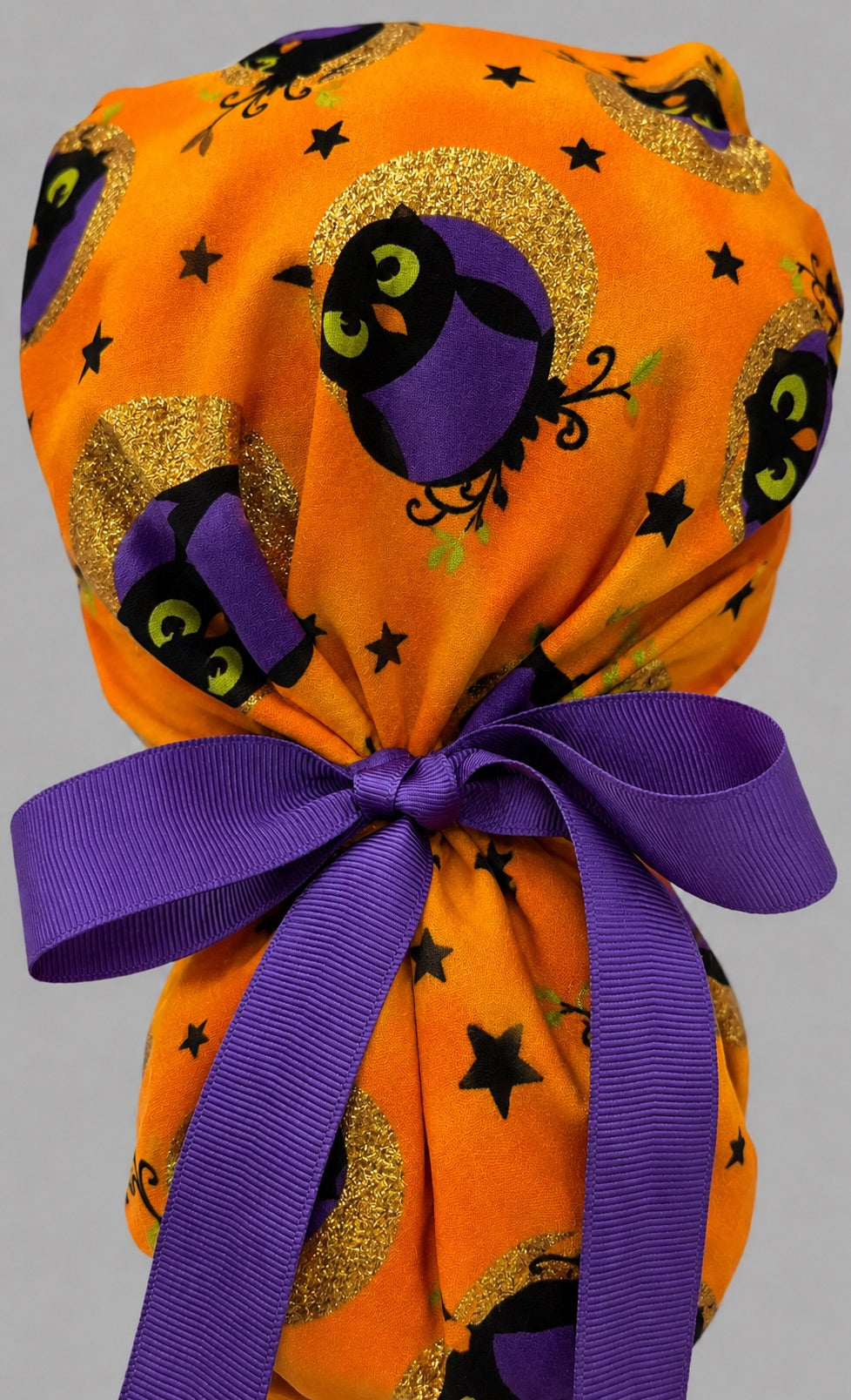 Woman wearing Halloween owl ponytail scrub cap, front view smiling with purple witch owls, gold moons and stars on orange with purple band and purple ribbon tie