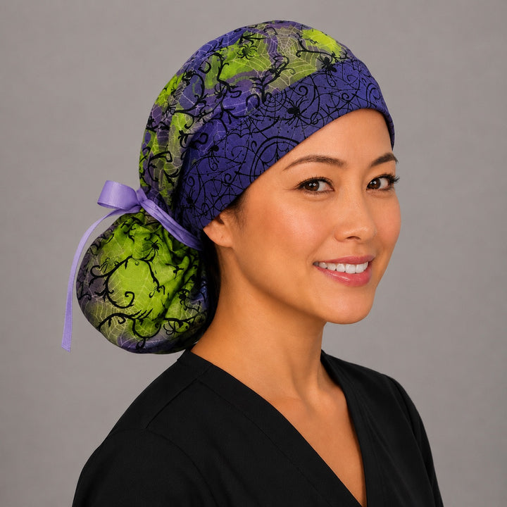 Woman wearing Halloween spider ponytail scrub cap, front view smiling with black spiders and webs on purple and lime green with purple ribbon tie