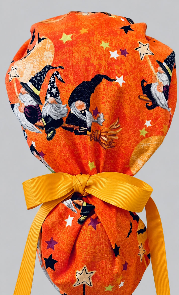 Woman wearing Halloween witch gnome ponytail scrub cap, front view smiling with witch gnomes, black cats and bats on orange with green spiderweb band and yellow ribbon tie