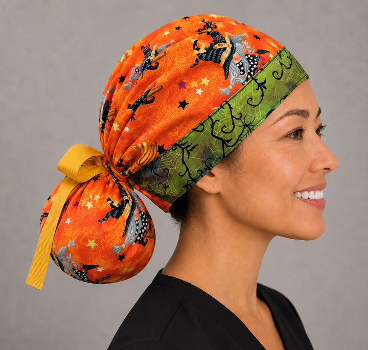 Halloween witch gnome ponytail nurse cap left three-quarter angle on smiling model showing witch gnomes on broomsticks and stars on orange with green spiderweb band and yellow ribbon