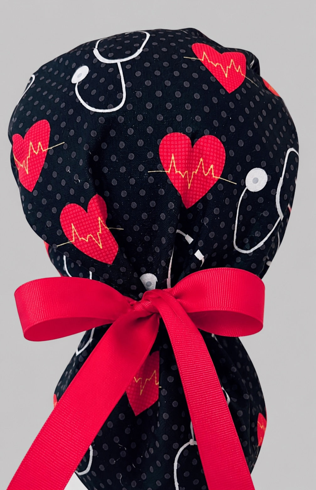 Heartbeat EKG cardiology ponytail scrub cap back view close-up showing gathered pouch of red hearts, gold EKG lines and white stethoscopes on black with red ribbon bow