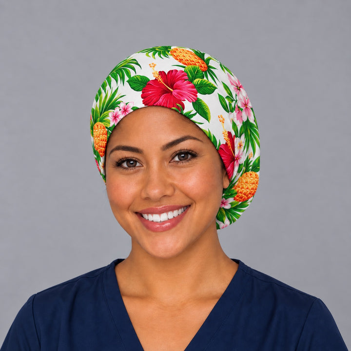 Woman wearing a Hawaiian hibiscus and pineapple Euro scrub cap, front view — pink blooms and pineapples on white, adjustable tropical surgical hat