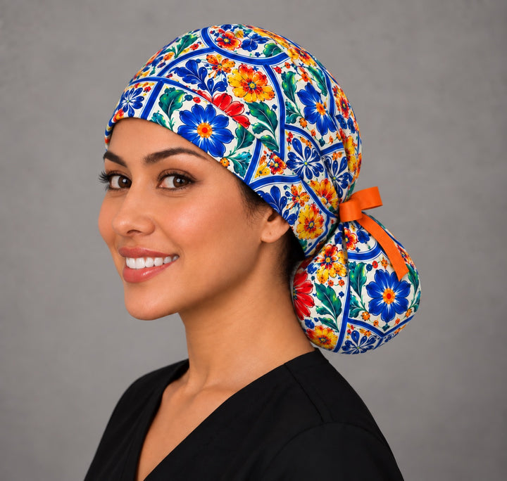 Left three-quarter angle of a smiling woman in a Hispanic heritage ponytail scrub hat, with blue, red and yellow blooms wrapping the side and a short orange bow