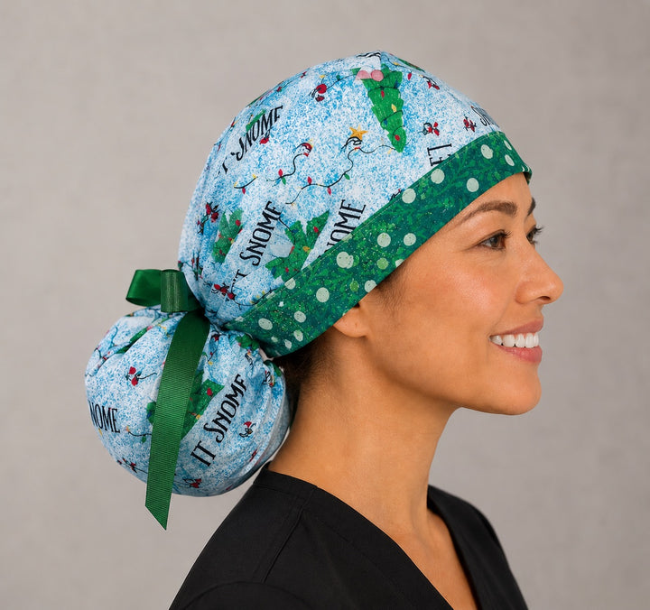 Woman wearing Let it Snome Christmas gnomes ponytail scrub cap, front view smiling with tree-gnomes and Let It Snow text on sky blue with green snowball band and green ribbon tie