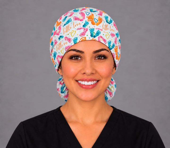 Smiling nurse in black scrubs wearing a Little Love baby feet ponytail scrub cap, front view with orange "little love" script and teal and pink footprints on the band