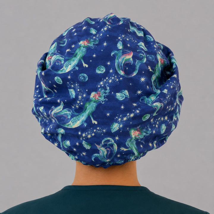 Mermaid ocean galaxy bouffant scrub cap back view showing full print of glowing mermaids, shells and stars on deep blue starry sea, OR World surgical hat