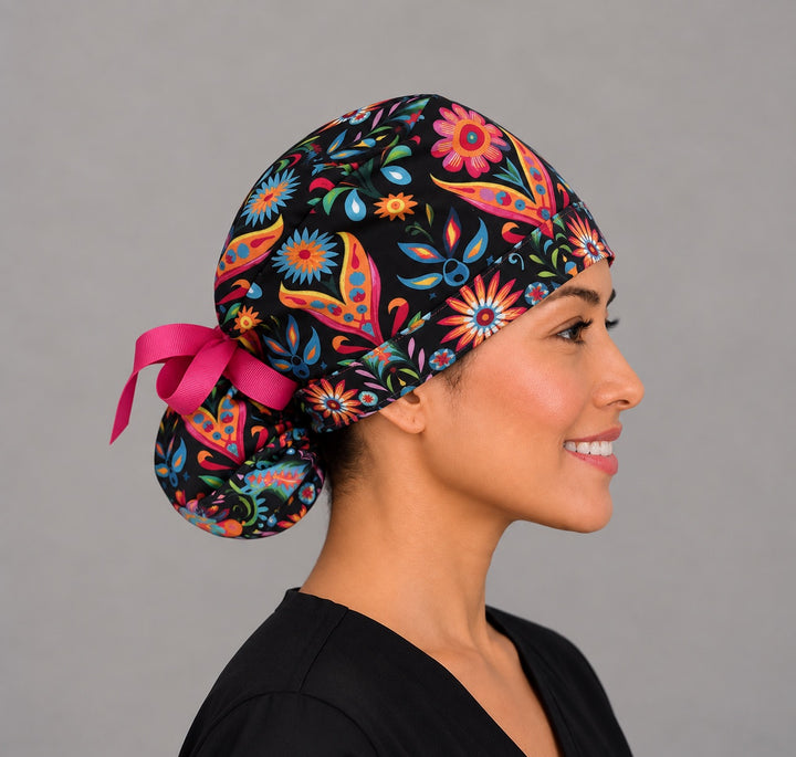 Woman wearing a Mexican floral ponytail scrub cap, front view — bright folk-art flowers on black with pink ribbon tie, adjustable surgical hat