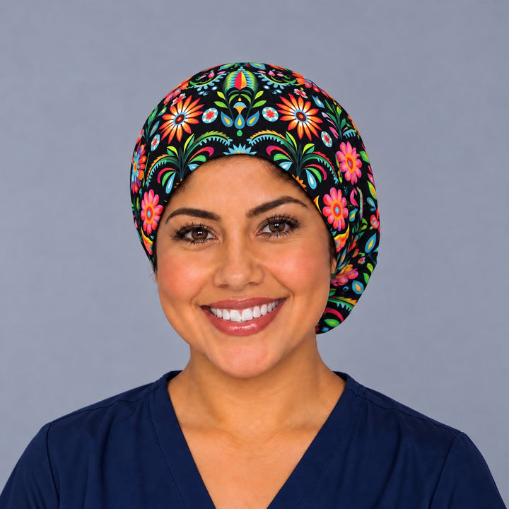 Smiling nurse in navy scrubs wearing a Mexican floral euro scrub cap, front view of the black folk flower band with orange sunbursts centered above the forehead