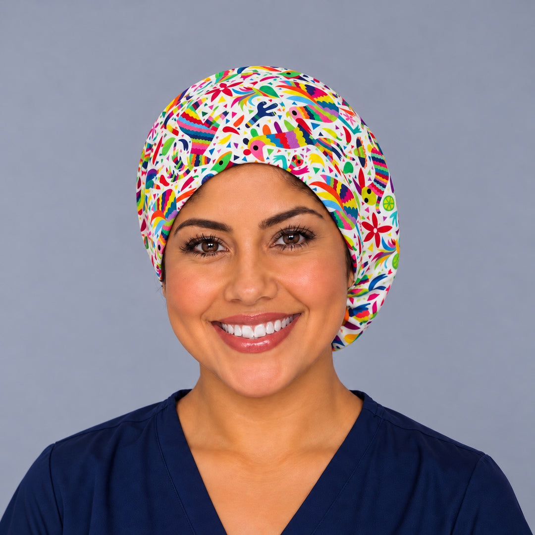 Smiling nurse in navy scrubs wearing a Mexican piñata euro scrub cap, front view of the white folk art band with rainbow piñata birds framing the face