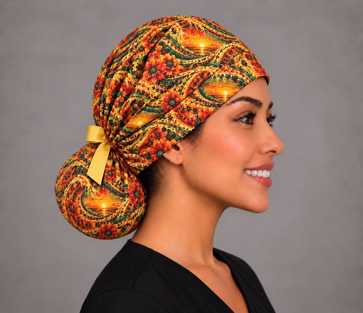 Woman wearing a Mexican sunset ponytail scrub cap, front view — gold waves, orange sunsets and red dahlias, handmade surgical hat for long hair