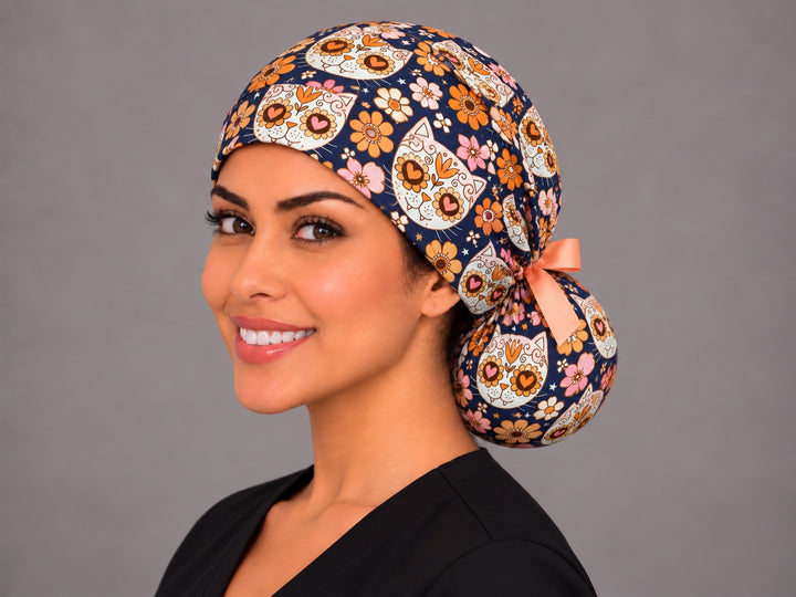 Three-quarter view of the navy Día de los Muertos cat ponytail nurse cap on a smiling OR nurse, low bun tucked in the printed pouch
