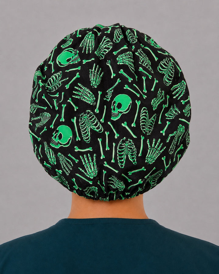 Woman wearing a neon green skeleton bouffant scrub cap, front view — glowing green skulls and bones on black, adjustable women&