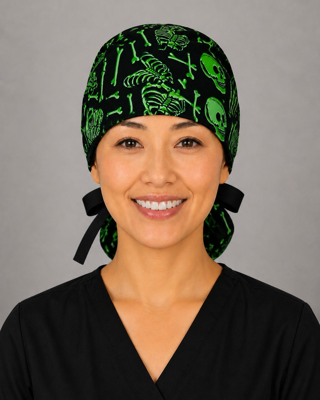 Woman wearing neon green skeleton ponytail scrub cap, front view smiling with glowing green skulls and bones on black and black ribbon tie